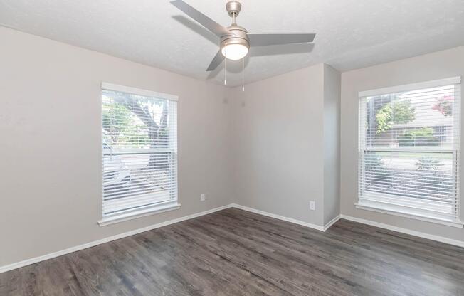 A light and airy room featuring two windows with blinds, a ceiling fan, and wooden flooring. The walls are painted in a neutral tone, and there are no visible furnishings, creating an open and spacious feel.