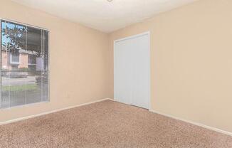 A clean, empty room with light beige walls and carpet. A large window with blinds allows natural light to enter, providing a view of the outdoors. On one wall, there is a white closet door. The space has a bright and open feel, suitable for various uses.