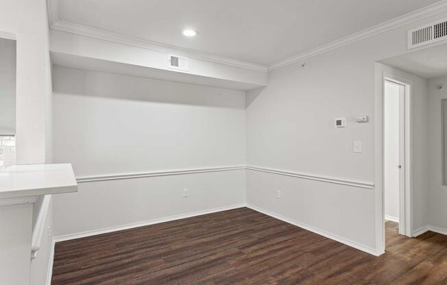 Empty room featuring light-colored walls, a wood-style floor, and crown molding. A small area with a wall-mounted thermostat is visible. There is a doorway leading to another room, and a counter space on the left side. Natural light comes from an overhead light fixture.