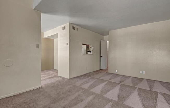 A spacious, empty living room here at High Plains with soft beige carpet, neutral walls, and an open layout, featuring a pass-through window to the kitchen and multiple wall openings that create a bright, flexible space for everyday living.