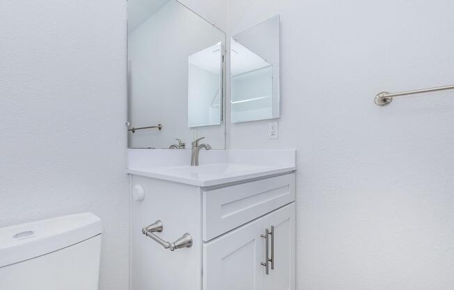 A clean, modern bathroom featuring a white vanity with a sink and a wide mirror above it. There is a toilet visible on the left side of the image. The walls are painted white, and the space appears bright and well-lit. A towel rack is mounted on the right wall.