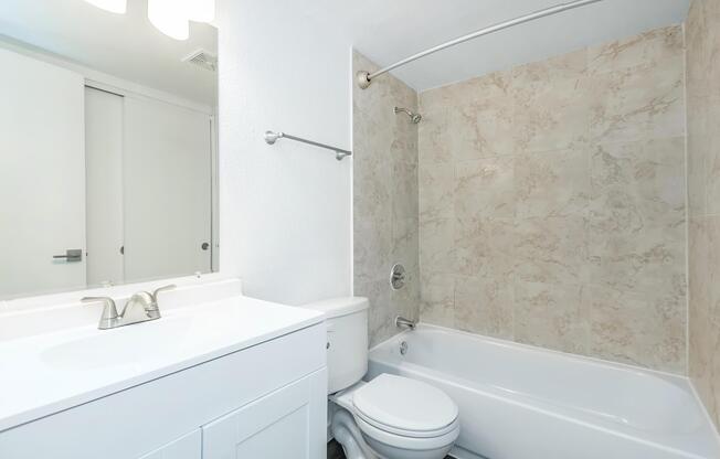 A clean, modern bathroom featuring a white bathtub and toilet. The walls are finished with light beige tiles, and there is a white vanity with a double sink. A large mirror is above the vanity, and there is a curved shower rod above the tub. The overall ambiance is bright and minimalist.