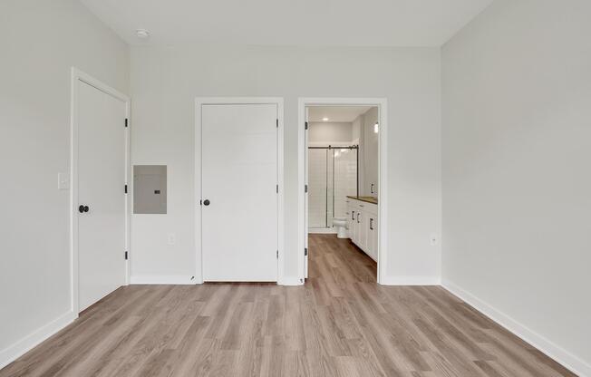 A spacious, light-colored room featuring a neutral wall, laminate flooring, and two white doors. One door leads to a bathroom, visible through an opening with modern fixtures. A mailbox slot is mounted on the wall, and the overall decor is minimalist and contemporary. Natural light enhances the open feel of the space.
