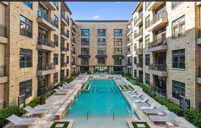 A swimming pool is surrounded by lounge chairs in a courtyard surrounded by apartment buildings.