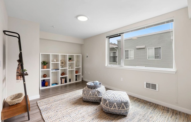 Cozy Living Area with Plank Flooring, Spacious Cozy Rugs, White Bookshelving, a large window and two stools in the middle of the room at Arabella Apartment Homes, Washington, 98155