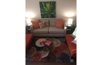 A cozy living room scene featuring a light gray sofa with orange accent pillows, flanked by two orange chairs. A circular coffee table holds a red book, and a colorful patterned area rug covers the floor. Two lamps illuminate the space, and a green plant artwork hangs on the wall, adding a touch of decor.