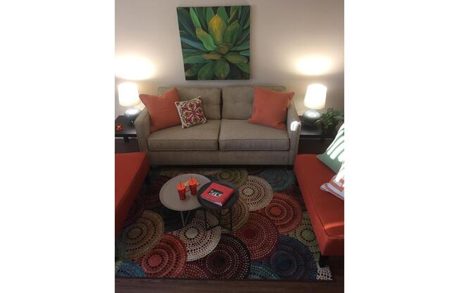 A cozy living room scene featuring a light gray sofa with orange accent pillows, flanked by two orange chairs. A circular coffee table holds a red book, and a colorful patterned area rug covers the floor. Two lamps illuminate the space, and a green plant artwork hangs on the wall, adding a touch of decor.