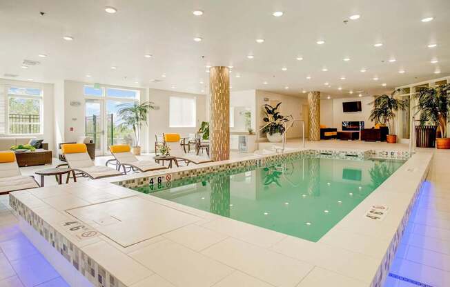 A resort-style indoor pool here at Heritage Hills featuring clear turquoise water, a tiled pool deck, and stainless handrails for easy access. The space includes cushioned lounge chairs with yellow accent pillows, patterned columns, potted greenery, and ample recessed lighting, creating a bright, relaxing environment designed for comfortable year-round use.