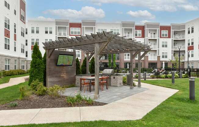 A wooden pavilion with a table and chairs is in the middle of a grassy area in front of apartment buildings.