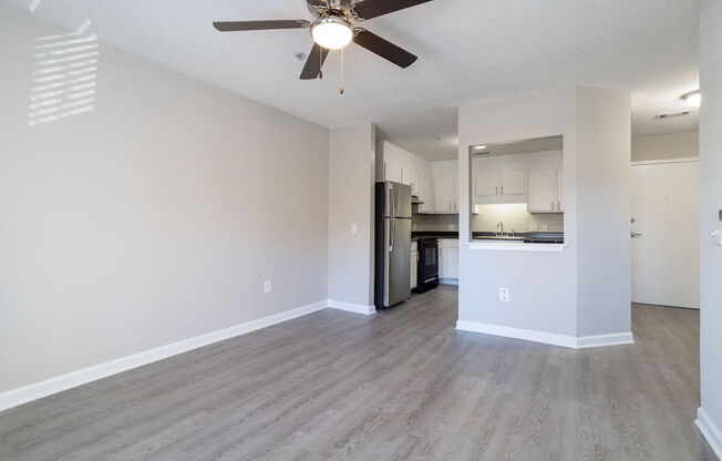 A spacious room with a ceiling fan and light fixture, hardwood flooring, and a kitchen area with stainless steel appliances.