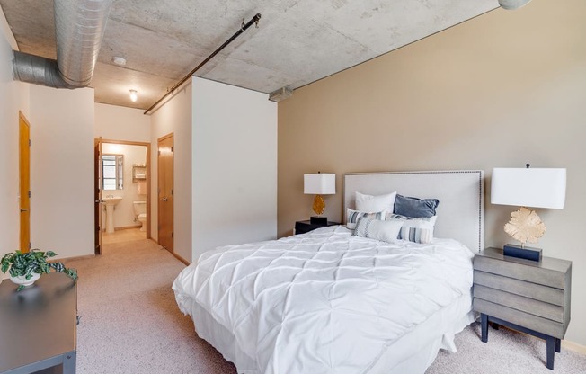 Bedroom with white comforter and two lamps on either side of bed and the bathroom down the hall