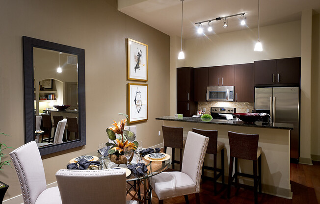 An open-concept kitchen and dining space with a four-person table, white walls, a taupe accent wall, dark wood cabinets, and stainless appliances.