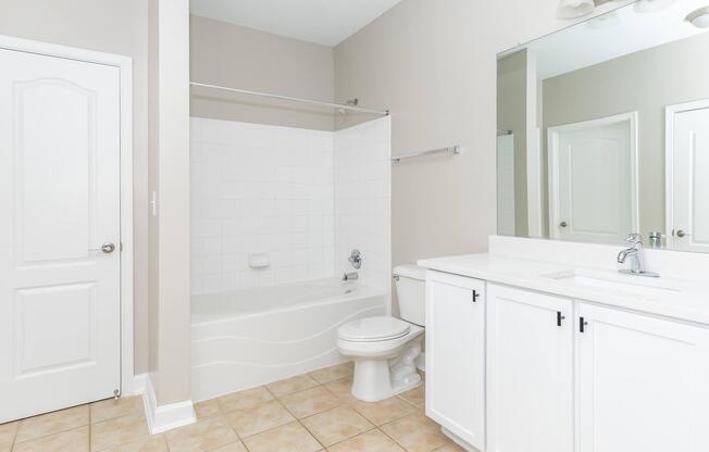 Modern bathroom featuring a bathtub with a shower curtain, a toilet, and a vanity with a sink. The walls are painted light beige, and there is a large mirror above the sink. The floor is tiled with light-colored tiles, creating a clean and spacious look. A door leads to another room.