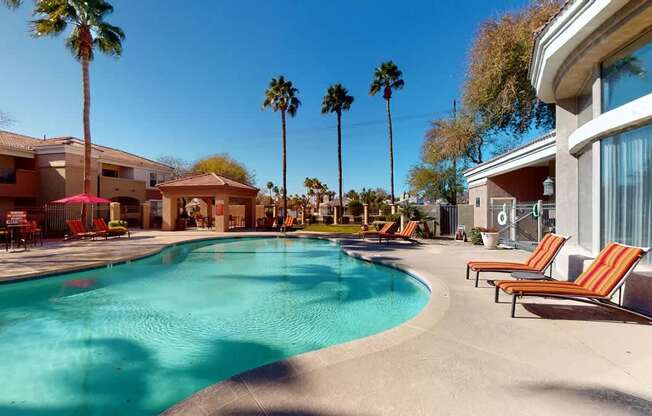 A sunlit resort-style pool area here at Dobson Towne Center showcasing a sparkling curved swimming pool, striped lounge chairs, and a spacious deck perfect for relaxing under the Arizona sky. Tall palm trees, shaded seating areas, and nearby covered cabanas create a tranquil atmosphere ideal for enjoying warm afternoons and gathering with neighbors.