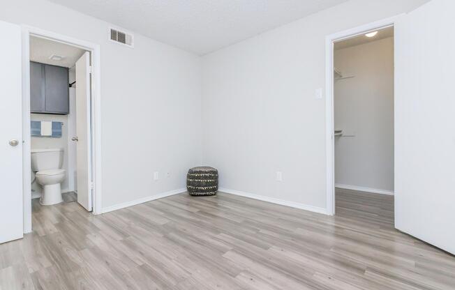 A clean, minimalist room with light gray walls and wood-like flooring. Two doors are visible: one leading to a bathroom with a toilet and the other to a closet. A small decorative stool is placed in the corner, adding a subtle touch to the empty space. Natural light brightens the room.