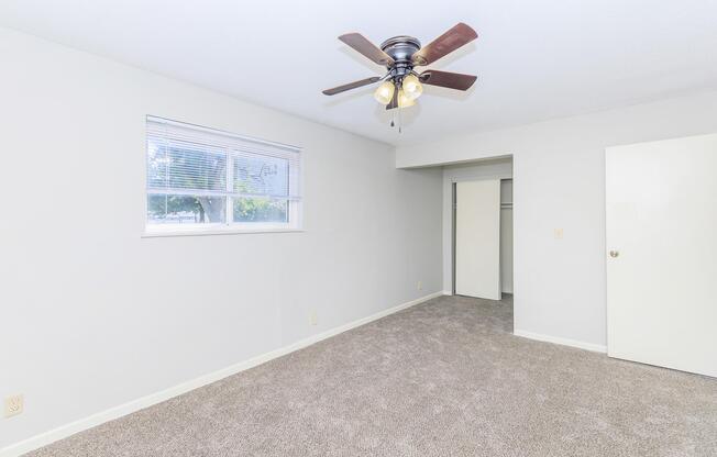 A spacious room with light-colored walls and carpeted flooring. A ceiling fan with a light fixture hangs from the ceiling. There is a window allowing natural light, and a doorway leading to a closet or additional space. The room is empty, creating a fresh and airy atmosphere.