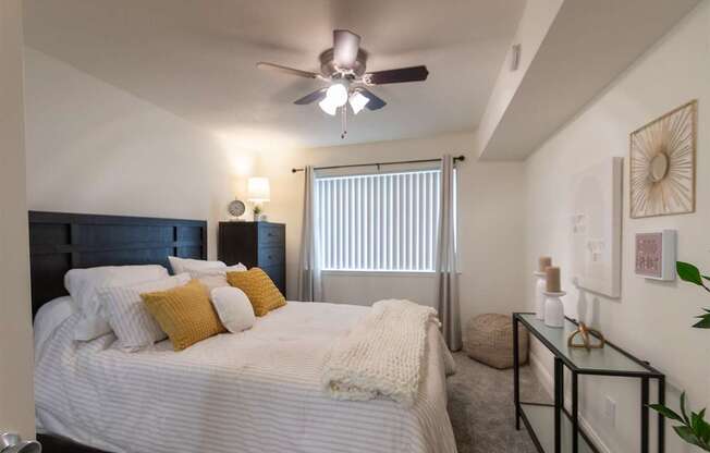 A bedroom with a bed, a ceiling fan, and a window with blinds in the 558 square foot 1 bedroom, 1 bath model apartment at The Summit at Midtown Apartments in the Dallas Midtown neighborhood of Dallas, TX.