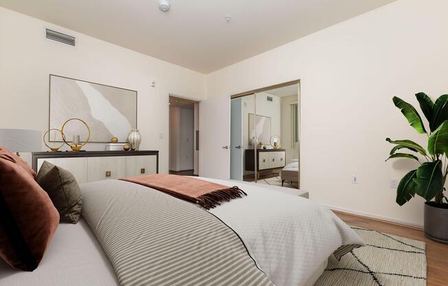 A modern bedroom featuring a neatly made bed with decorative pillows and a textured blanket, a stylish nightstand with decorative objects, a full-length mirror, and a leafy indoor plant in the corner. Light-colored walls enhance the spacious feel, and thereâs a doorway leading to another room visible in the background.