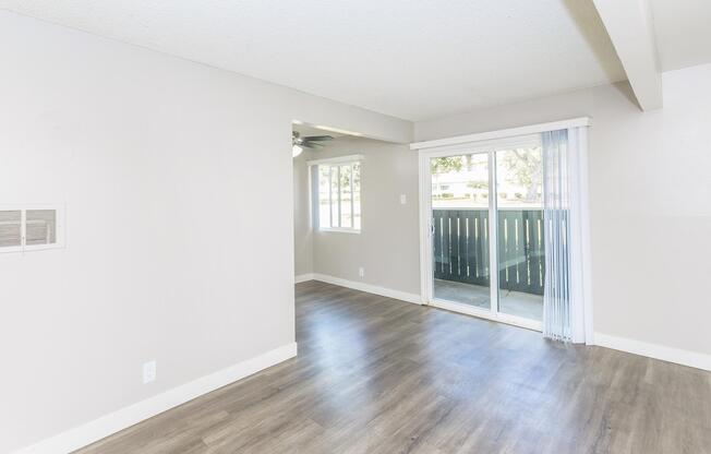 A spacious, light-filled living room with neutral walls and flooring. A sliding glass door leads to a balcony or outdoor area, and a window allows natural light. The room features a ceiling fan and is empty, creating a clean, inviting atmosphere.