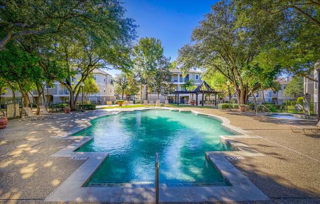 Sparkling swimming pool surrounded by a sun deck with lounge chairs and mature trees at Saxony at Chase Oaks Apartments in Dallas, TX. The area includes a shaded pergola and is framed by residential apartment buildings in the background.