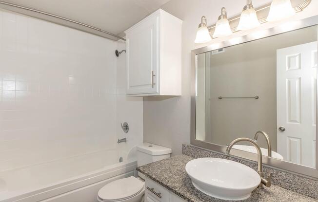 A modern bathroom featuring a white bathtub, a granite countertop with a round sink, a toilet, and a mirrored cabinet with overhead lighting. The walls are painted in a light color, and there is a towel bar next to the doorway. The overall design is clean and contemporary.