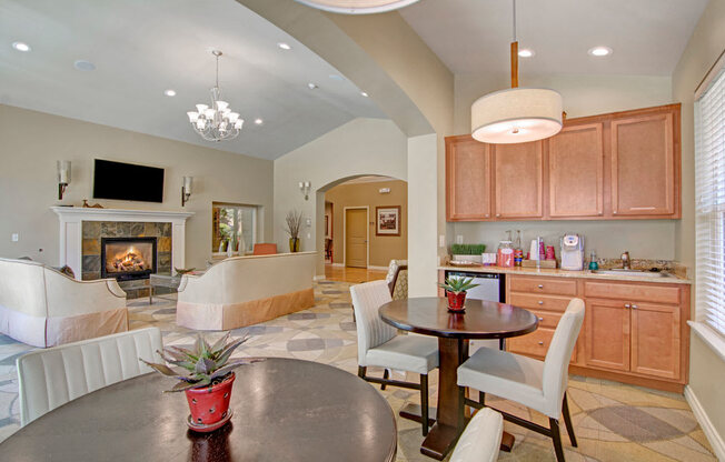 A spacious community lounge here at Asbury Park showcasing a comfortable seating area arranged around a tiled fireplace with a decorative mantel and wall-mounted television, complemented by a kitchenette with wood-style cabinets, countertop appliances, pendant lighting, café-style tables and chairs, soft neutral walls, patterned flooring, and an open layout designed for relaxing and gathering.
