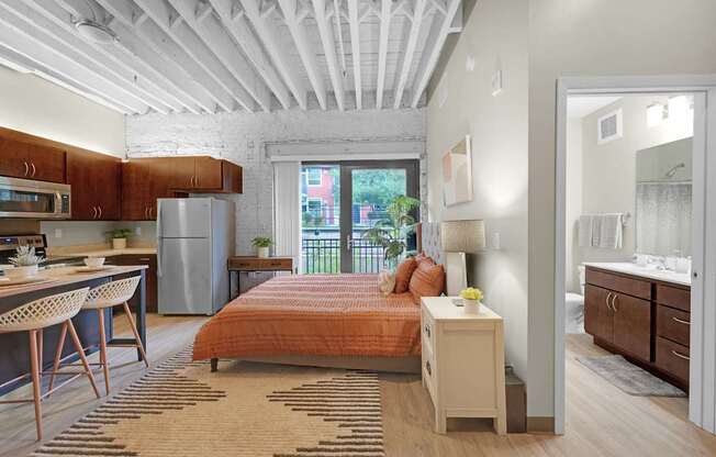 A bedroom with a bed, nightstand, and a kitchen area with a refrigerator and microwave.