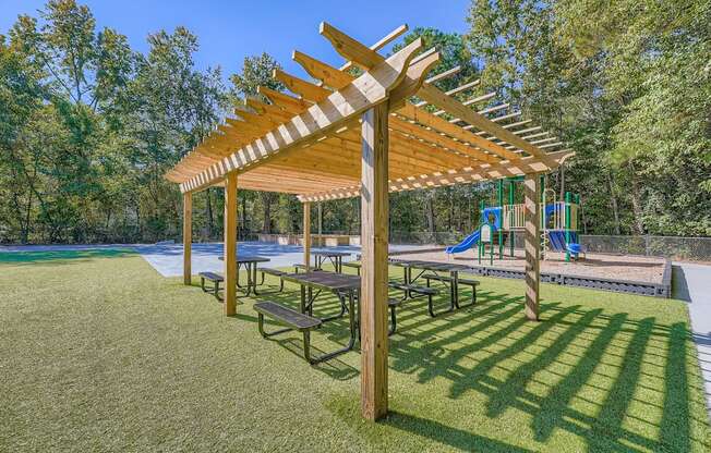 A wooden pergola with benches is situated in a grassy area with a playground in the background.