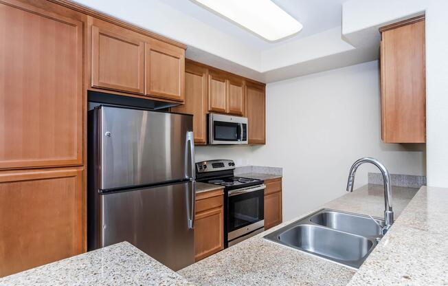 A modern kitchen featuring wooden cabinets, stainless steel appliances including a refrigerator, microwave, and oven. The countertop is made of speckled granite, with a double sink and a faucet. The kitchen is well-lit with a ceiling light and has a clean, contemporary design.