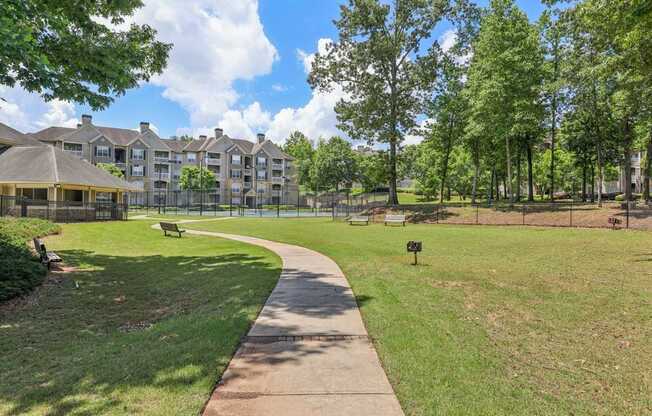 A walkway leads through a grassy area with grills,  trees and apartment buildings in the background.