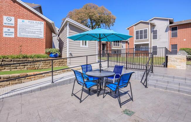 A patio area featuring a round table with four blue chairs under a large turquoise umbrella. In the background, several residential buildings and a sign displaying pool rules are visible. The setting is bright and sunny, with a clear blue sky.