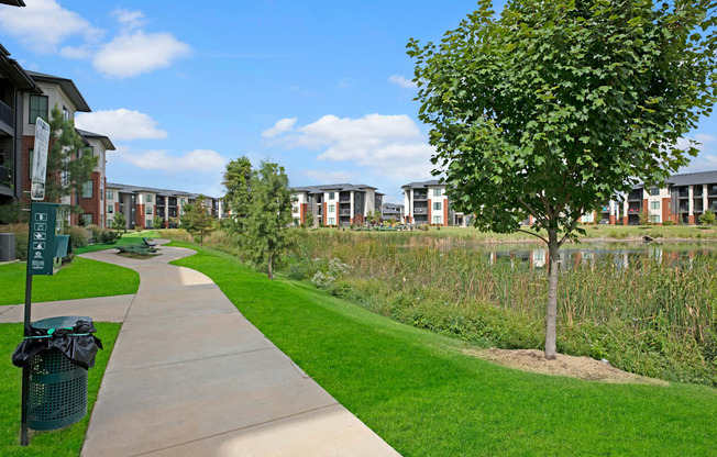 Paved path curves through a green park, lined with trees near 49 West Apartment buildings. Bright blue sky with clouds creates a serene and inviting atmosphere.