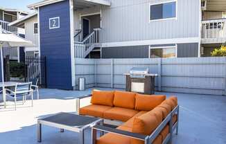 Outdoor lounge with umbrella-covered tables, gas BBQ, and an oversized orange patio couch and coffee table.at The Lakes Apartments, Moses Lake, WA