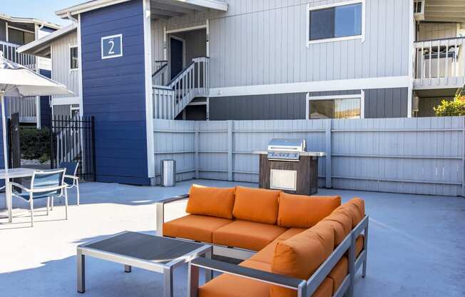 Outdoor lounge with umbrella-covered tables, gas BBQ, and an oversized orange patio couch and coffee table.at The Lakes Apartments, Moses Lake, WA