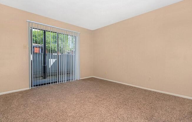 A vacant room with beige walls and carpeted flooring. There are vertical blinds covering a sliding glass door that leads to an outdoor area. The space is well-lit with natural light coming through the blinds, providing a clean and simple aesthetic.
