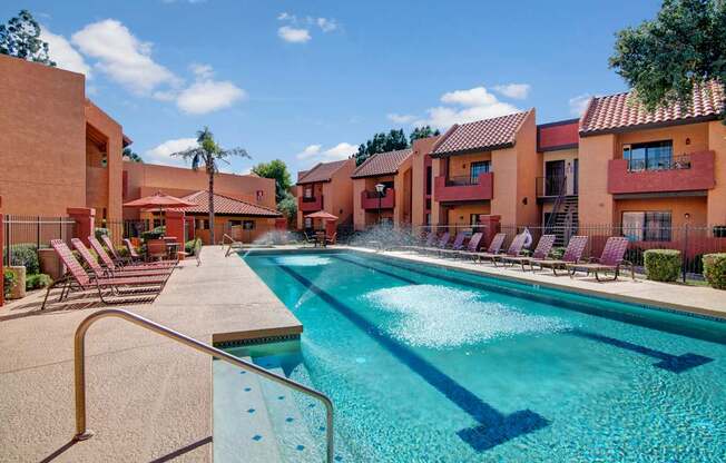 A sunlit resort-style pool here at Aspire Pinnacle Park featuring a sparkling lap pool with built-in lane markers, surrounding lounge chairs, shaded seating areas, and red stucco buildings with tile roofs. Palm trees and landscaped courtyards create a warm, inviting atmosphere perfect for relaxing, swimming, or enjoying Arizona sunshine.