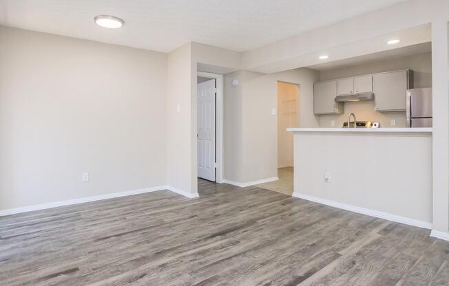 A spacious, well-lit living area with light-colored walls and wooden-style flooring. A small white bar separates the living space from a kitchen in the background. There's a door leading to another room, and the overall atmosphere is clean and modern. 