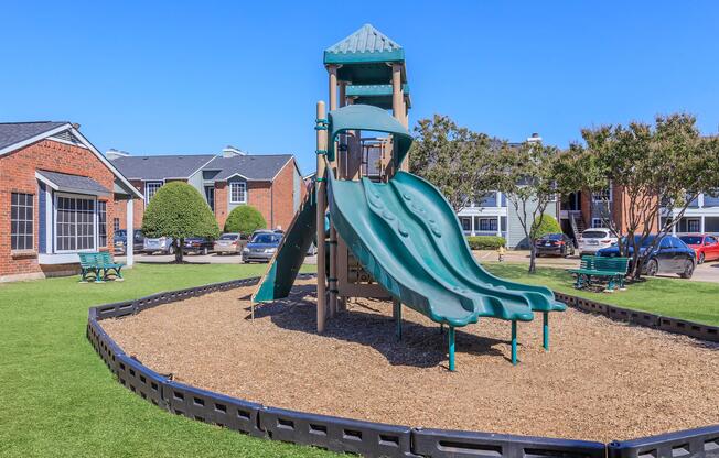 A playground featuring a large green slide structure on a bed of mulch, surrounded by a grassy area. Nearby, there are benches and parked cars, with apartment buildings visible in the background under a clear blue sky.