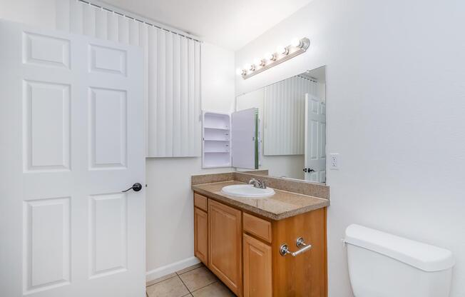 A bright bathroom featuring a wooden vanity with a sink, a mirror above the sink, a light fixture, and a wall-mounted cabinet. The room has white walls, a tiled floor, a toilet on the right, and a door leading to another area. A window is covered with vertical blinds.