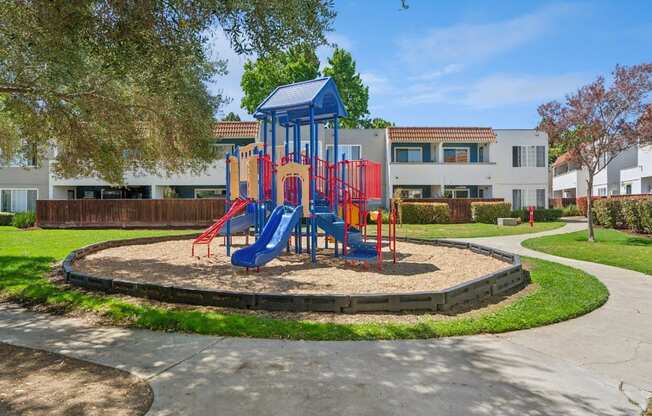 A playground with a blue and red slide in the middle of a grassy area.