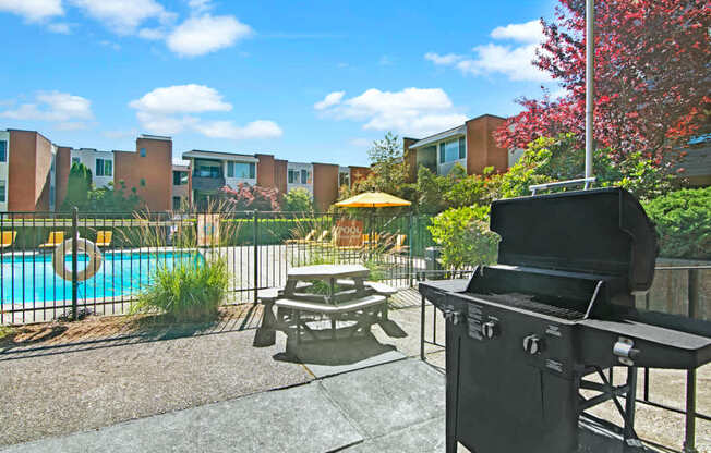 A welcoming outdoor grilling area here at Watercrest with a barbecue grill, picnic table seating, and views of the community swimming pool, creating a perfect space for relaxing and outdoor gatherings.