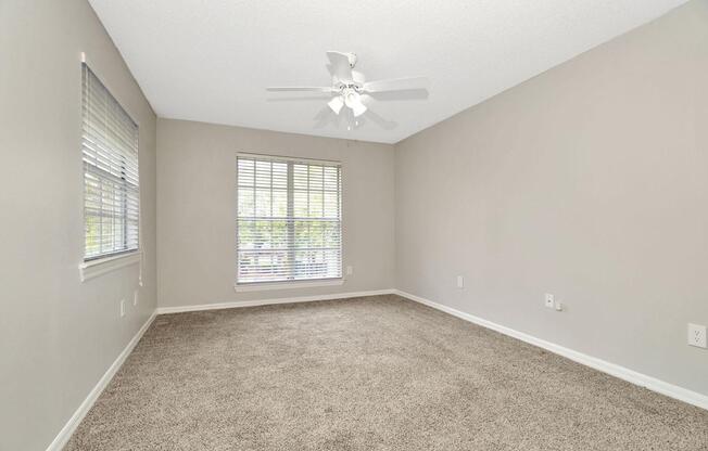 A spacious room with light beige walls and a ceiling fan. It has large windows with blinds, allowing natural light to enter. The floor is covered with plush, light brown carpet, and the space is empty, providing a clean, uncluttered appearance.