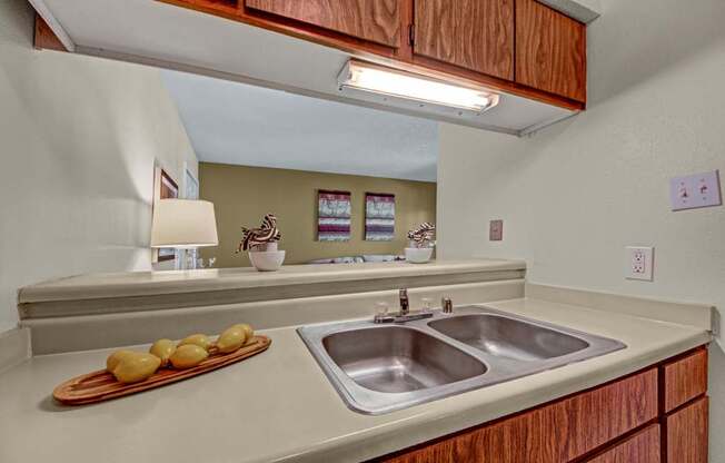 A functional kitchen here at Cypress Pointe with warm wood cabinets, beige countertops, and a double stainless steel sink beneath bright overhead lighting. An open pass-through overlooks the living area, creating a connected and practical layout for everyday use.