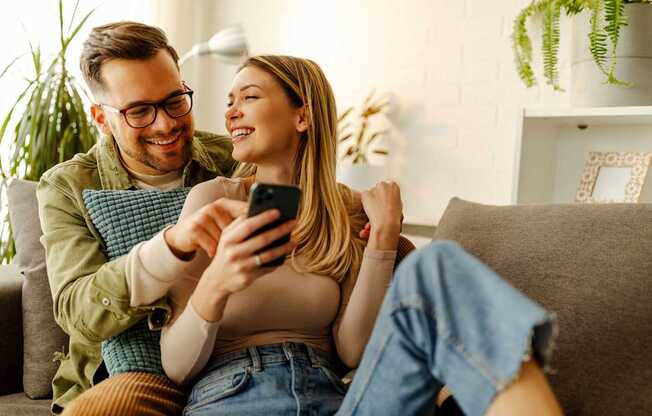 A man and a woman are sitting on a couch, looking at a smartphone together.