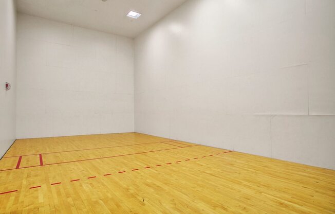 A spacious indoor racquetball court here at Allegro featuring smooth hardwood flooring with clearly marked red boundary lines, tall white walls, and bright overhead lighting, creating a clean and open recreational space designed for active play, solo practice, or friendly matches in a private indoor setting.