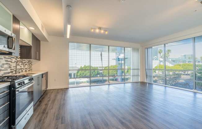 an empty living room with a large window and a kitchen with stainless steel appliances