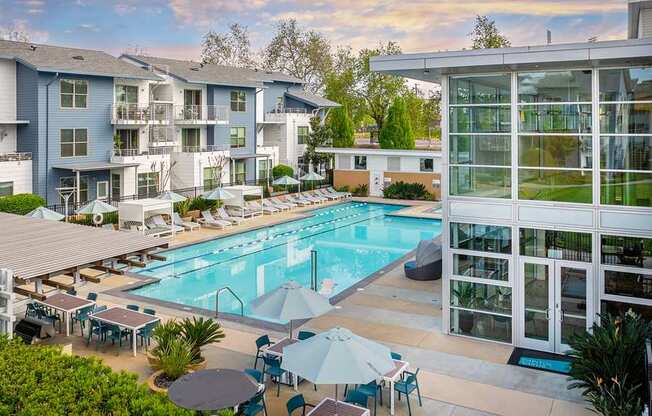 A large swimming pool surrounded by lounge chairs and umbrellas in front of apartment buildings.