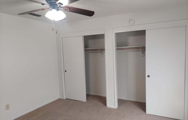Interior view of a room featuring a ceiling fan with light, two sliding closet doors, and beige carpet. The walls are painted white and the closets are empty, showcasing hanging rods without any items inside.