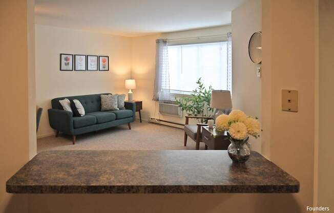 A living room with a dark green couch, a coffee table with a vase of flowers, and a window with white curtains.