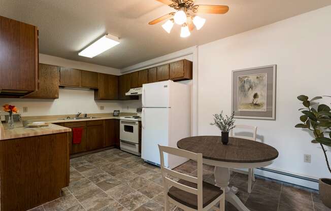 Bismarck, ND Rosser Apartments. A dining room with a table and two chairs. The overhead fan and light brighten the room. A kitchen with a white refrigerator freezer next to a white stove top oven are in the background